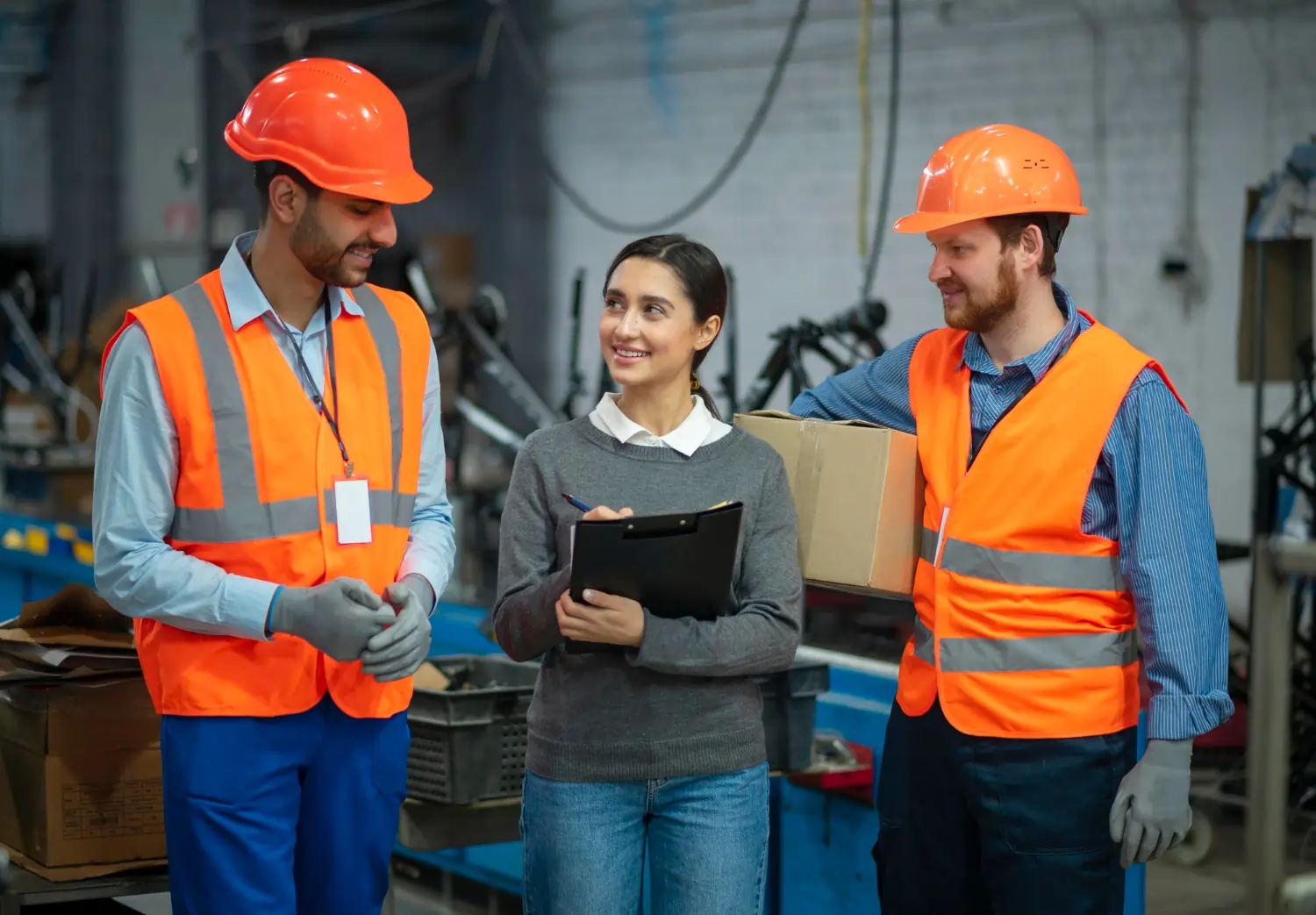 A smiling female supervisor holding a clipboard reviews operational data with two factory workers wearing bright orange safety vests, hard hats, and protective gloves on an active manufacturing floor.
