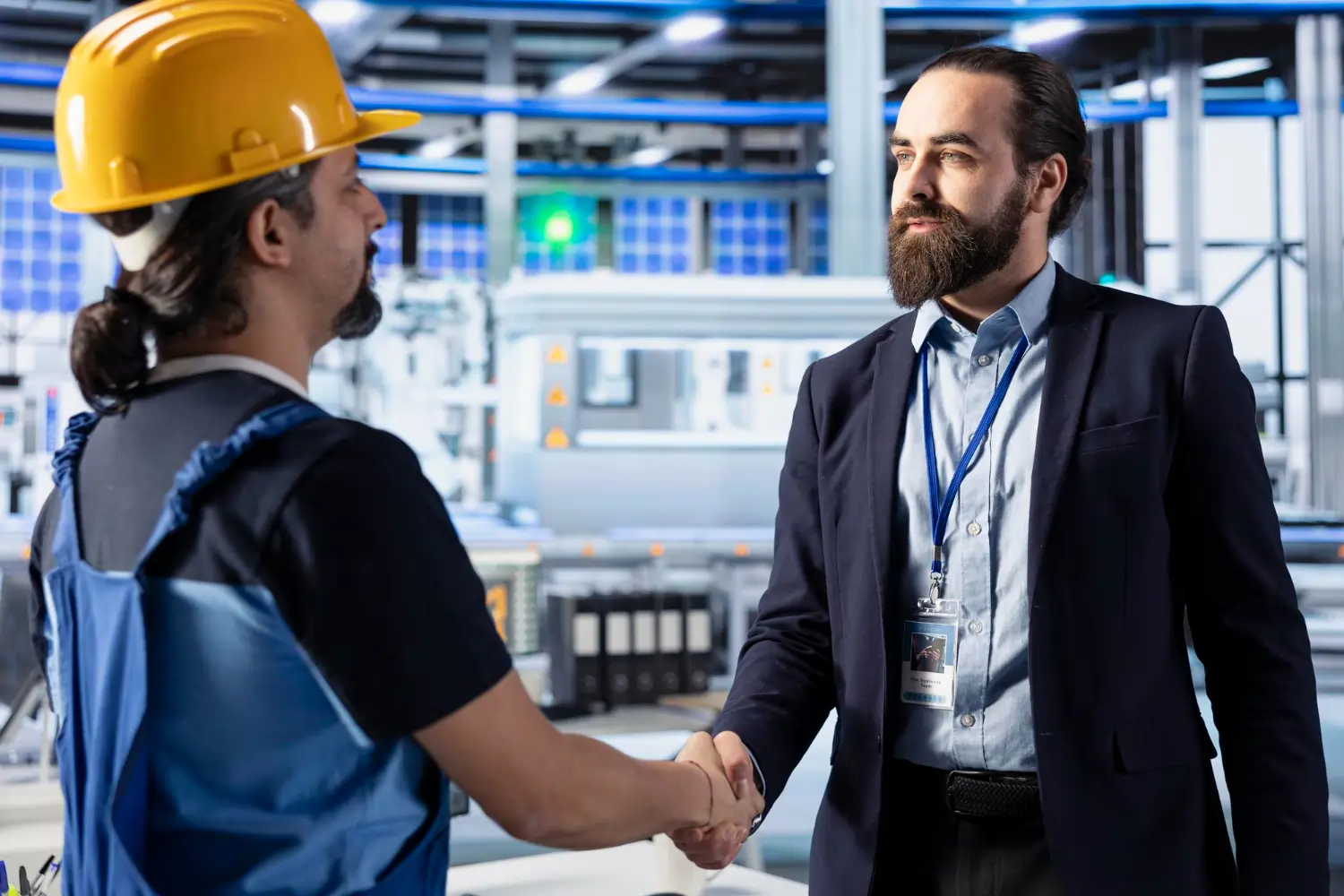 A professional corporate manager with a beard and lanyard shakes hands with a factory worker wearing a yellow hard hat and blue overalls, sealing a successful compliance agreement on a high-tech manufacturing floor.